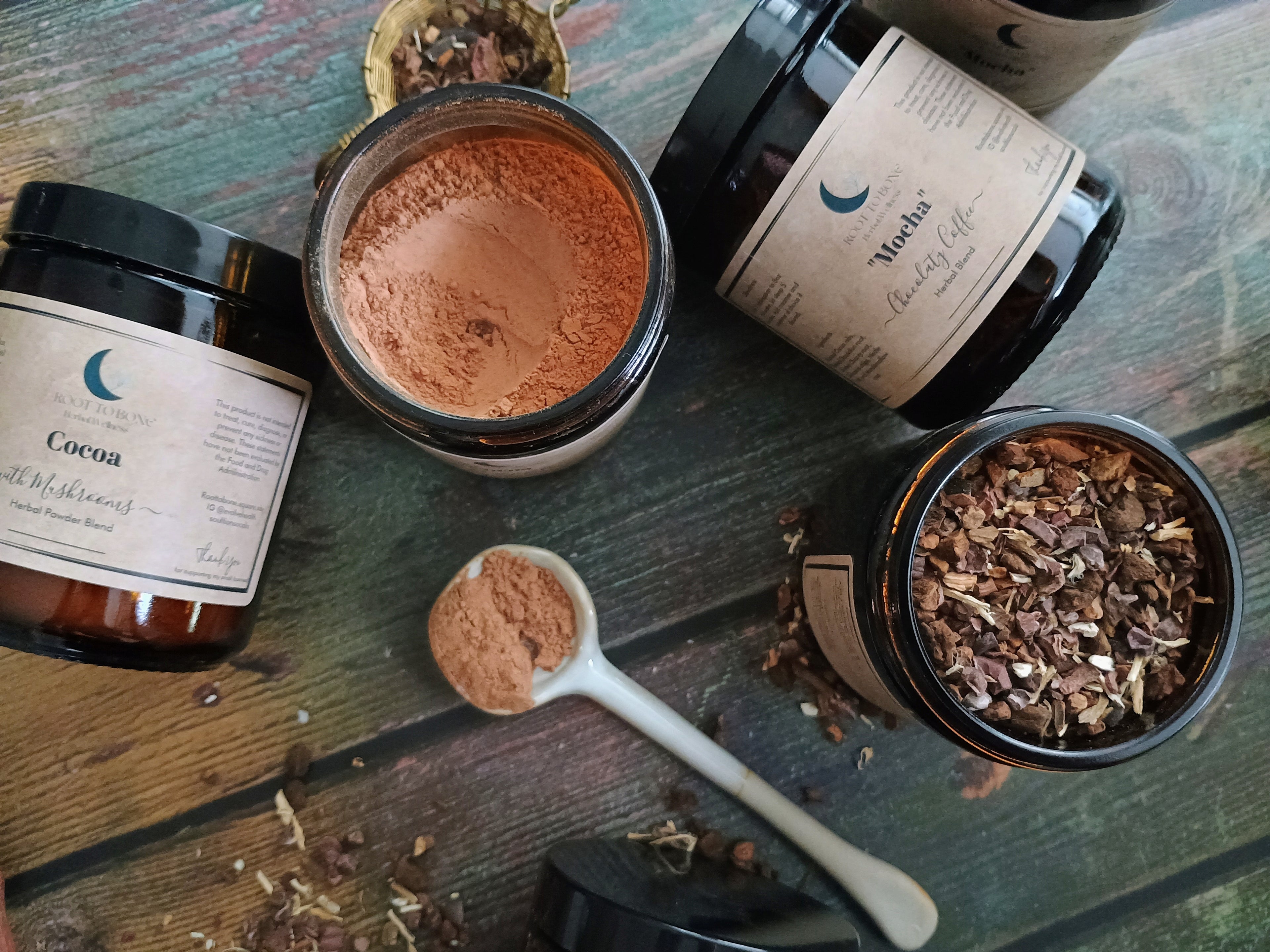 Jars of cocoa powder and dried herbs on a wooden surface with a spoonful of cocoa powder.