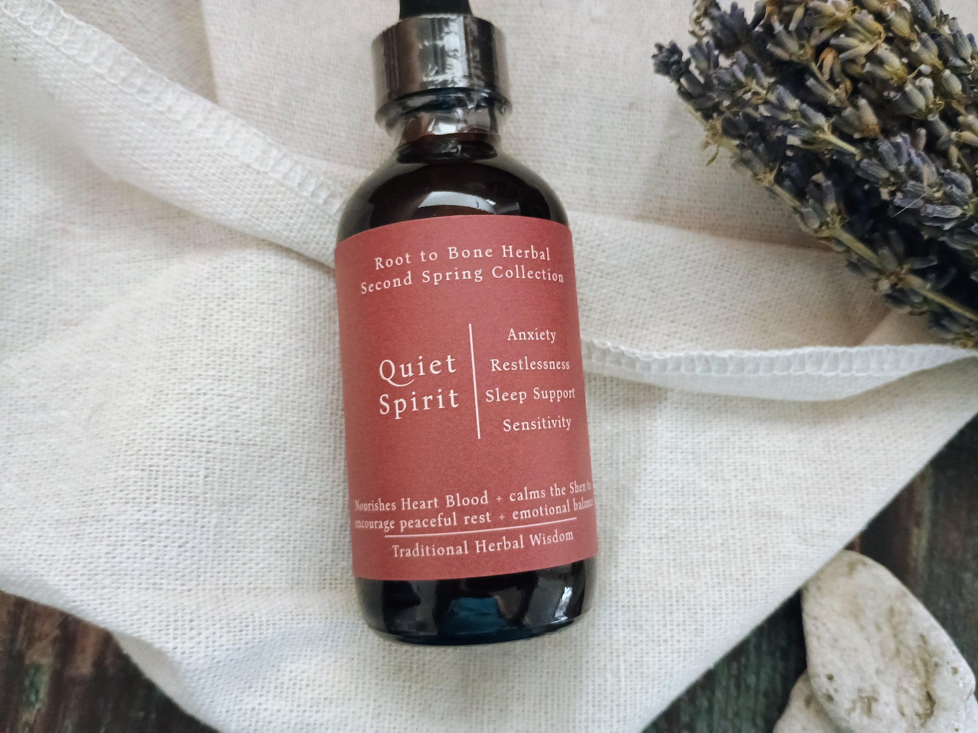 A dark amber glass dropper bottle labeled 'Quiet Spirit' by Root to Bone Herbal, resting on a textured white linen cloth with dried lavender sprigs in the background.