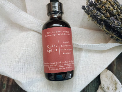 A dark amber glass dropper bottle labeled 'Quiet Spirit' by Root to Bone Herbal, resting on a textured white linen cloth with dried lavender sprigs in the background.