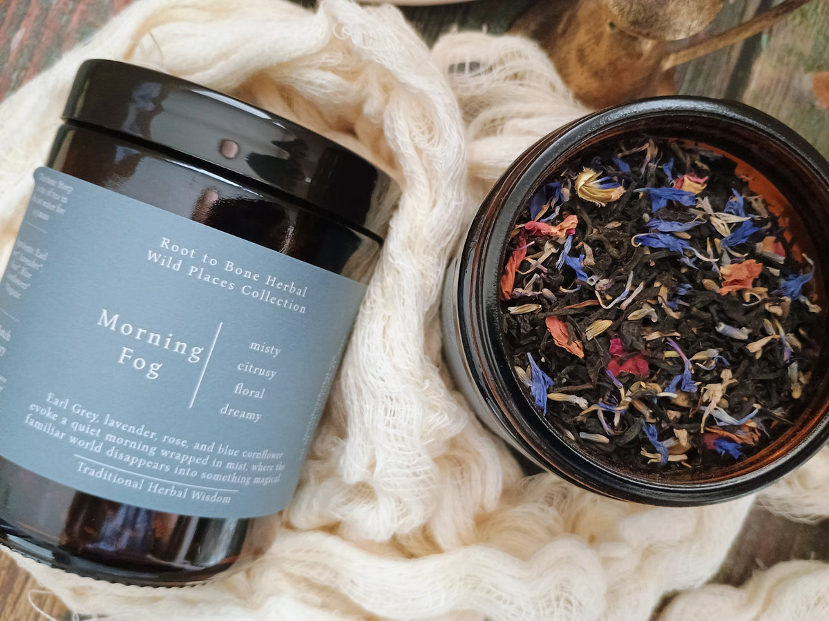 An open dark amber jar of Morning Fog herbal tea from Root to Bone Herbal Wild Flowers Collective, shown next to its grey label on a rustic wooden surface wrapped in cream-colored knit fabric.