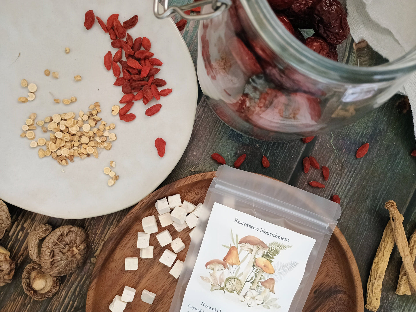 A clear stand-up pouch labeled 'Restorative Nourishment' sits on a wooden tray beside sliced white mushrooms, scattered red goji berries, and a glass jar filled with dried red fruits, arranged on a rustic dark wood surface.