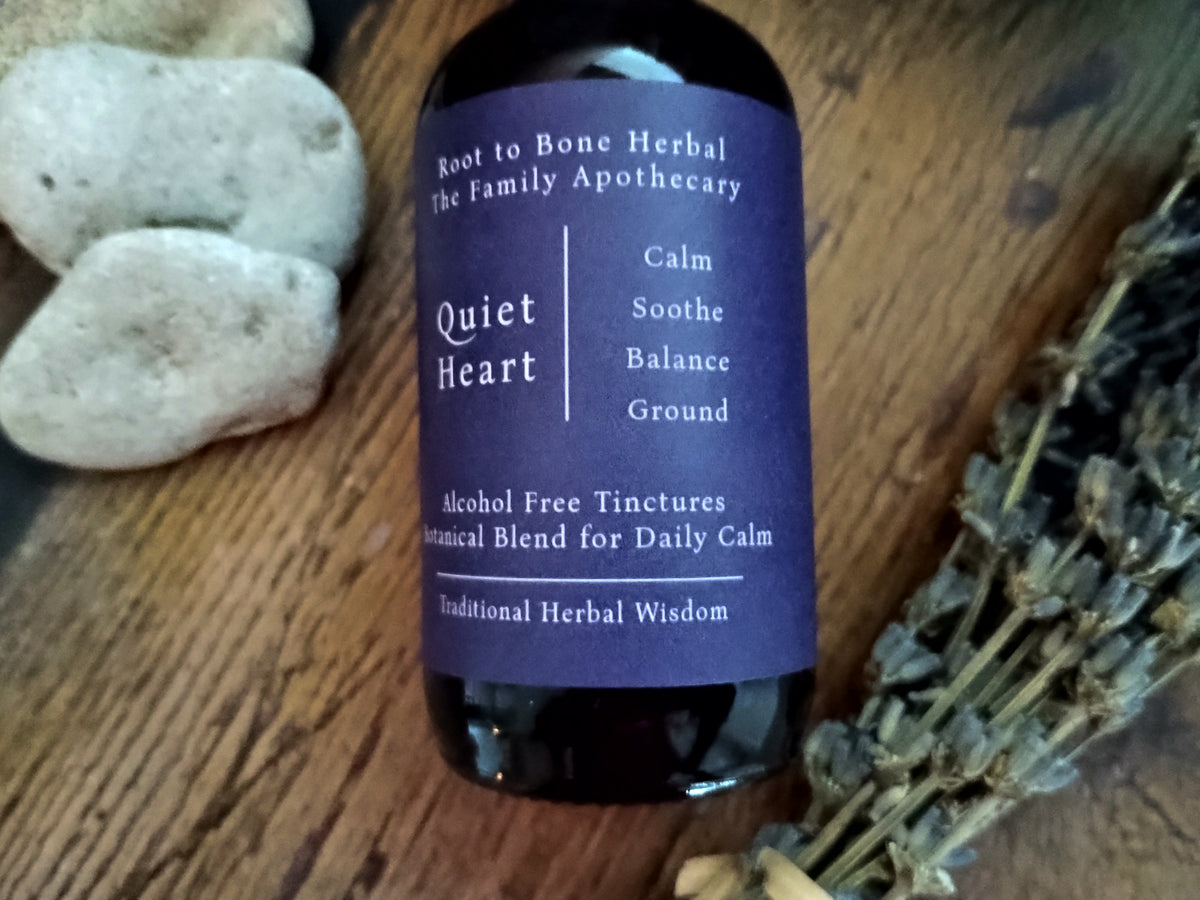 A dark glass bottle of Root to Bone Herbal The Family Apothecary 'Quiet Heart' tinctures, featuring a dark purple label, set on a rustic wooden surface with decorative stones and dried lavender.