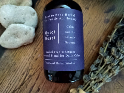 A dark glass bottle of Root to Bone Herbal The Family Apothecary 'Quiet Heart' tinctures, featuring a dark purple label, set on a rustic wooden surface with decorative stones and dried lavender.