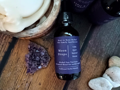 A dark amber glass dropper bottle labeled 'Moon Drops' by Root to Bone Herbal The Family Apothecary, styled on a rustic wooden surface with crystals and soap.