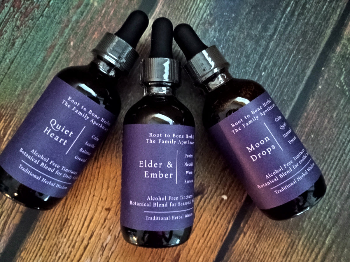 Three amber glass dropper bottles of Root to Bone Herbs homeopathic remedies arranged on a rustic wooden surface. The bottles feature purple labels for 'Quiet Heart', 'Elder & Ember', and 'Moon Drops'.