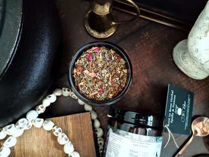 A top-down styled product shot of a herbal tea blend in a small black bowl, with a branded jar, spoon, beads, and wooden props on a dark wood surface. 