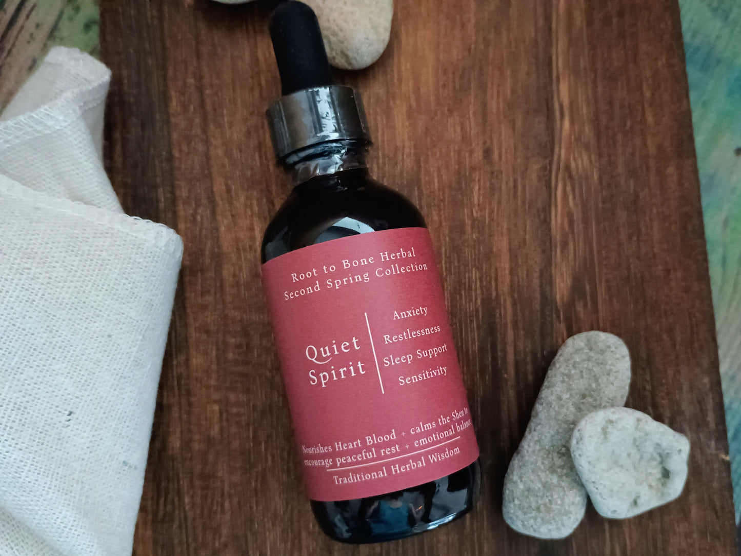 A dark amber glass dropper bottle labeled 'Quiet Spirit' by Root to Bone Herbal, placed on a rustic wooden surface with white stones and a white cloth nearby.
