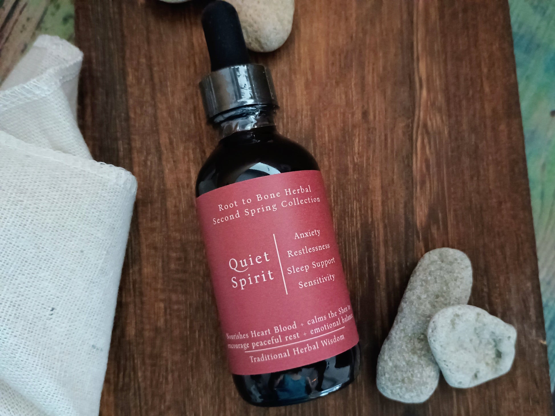 A dark amber glass dropper bottle labeled 'Quiet Spirit' by Root to Bone Herbal, placed on a rustic wooden surface with white stones and a white cloth nearby.