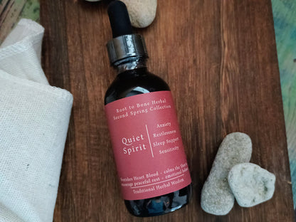 A dark amber glass dropper bottle labeled 'Quiet Spirit' by Root to Bone Herbal, placed on a rustic wooden surface with white stones and a white cloth nearby.