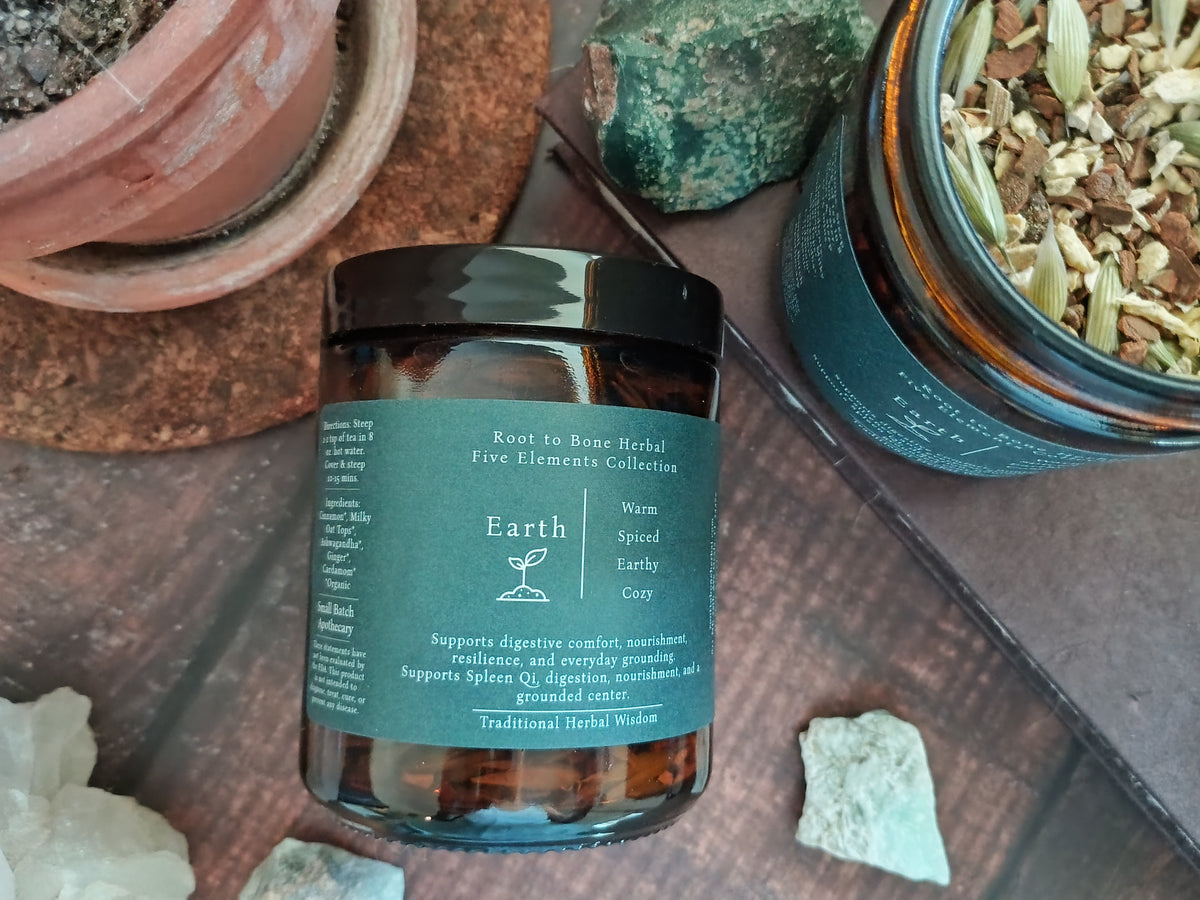 A glass jar of Root to Bone Herbal Five Elements Collection 'Earth' spiced and earthy herbal tea, styled on a rustic wooden surface with crystals, a green stone, and another open jar of dried herbs.