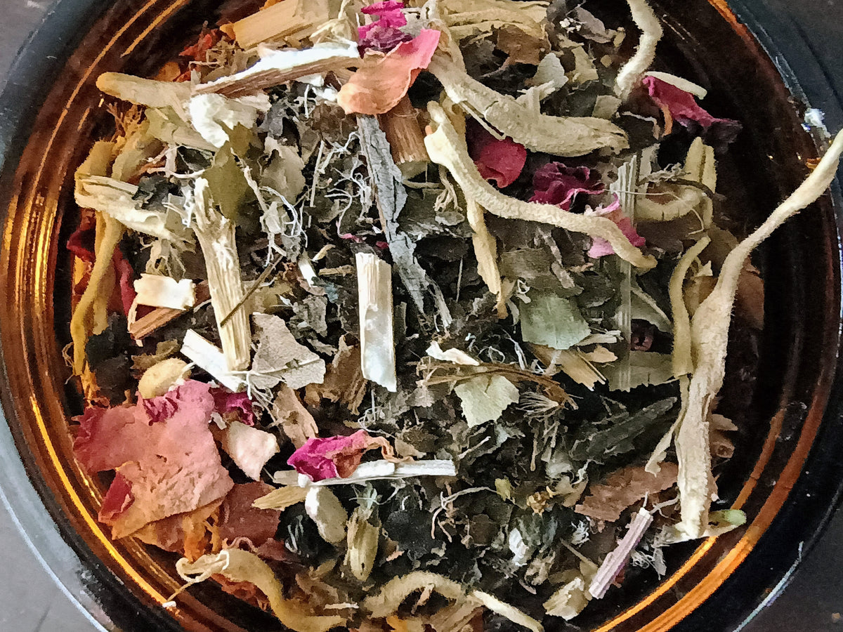 A close-up top-down view of a loose herbal tea blend inside a dark amber glass jar. The blend contains dried green leaves, pale beige roots or bark pieces, and pink dried flower petals.
