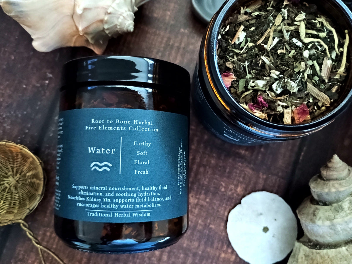 A dark amber glass jar labeled 'Water' from the Root to Bone Herbal Five Elements Collection, placed on a dark wooden surface next to an open jar showing loose herbal tea ingredients, with seashells and dried botanicals as props.