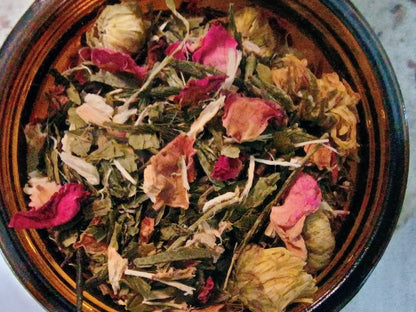 A top-down view of a dark brown ceramic bowl filled with loose herbal tea, featuring dried green leaves, pink rose petals, and pale yellow flower buds.
