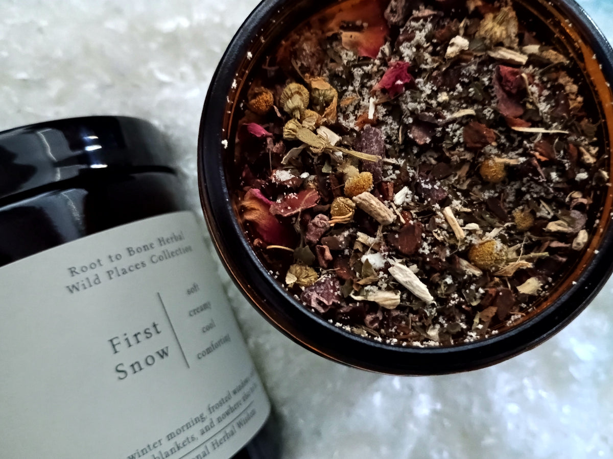 An open dark amber jar showing a dried herbal and rose petal blend inside, next to the black label reading 'First Snow' and 'Root to Bone Herbs Wild Places Collection' on a white textured background.