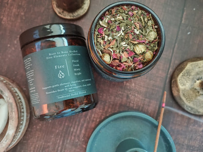 An amber glass jar labeled 'Roots to Bone Herbal Five Elements Collection Fire' shown next to an open jar filled with dried herbs and flowers, arranged on a rustic wooden surface with decorative stones and a teal incense holder.