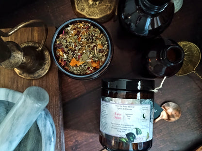 A jar of Root to Bone Herbal tea is displayed on a dark wooden table alongside a bowl of loose herbal tea, a marble mortar and pestle, and dark glass bottles.