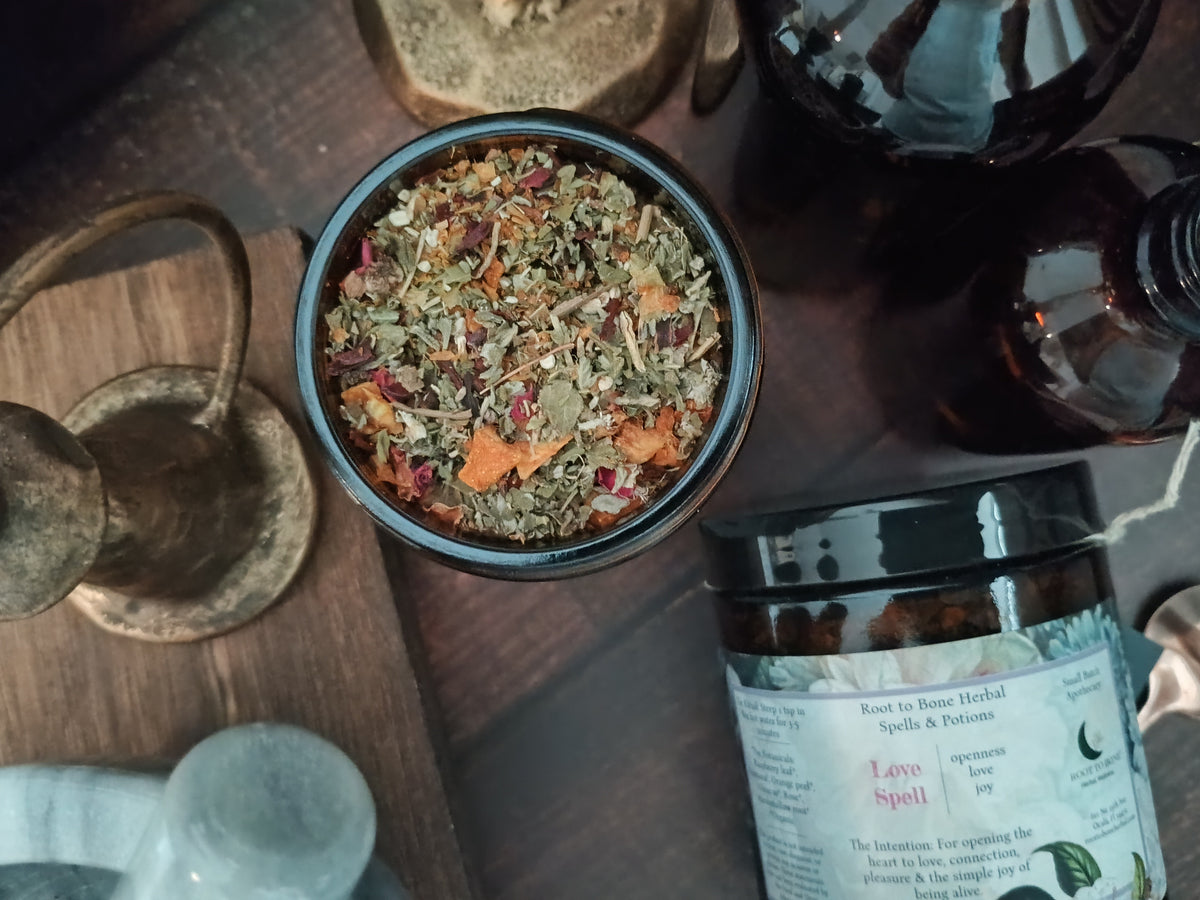 A top-down view of a dried herbal blend in a black bowl, alongside a jar labeled 'Love Spell' and dark amber bottles on a rustic wooden surface.