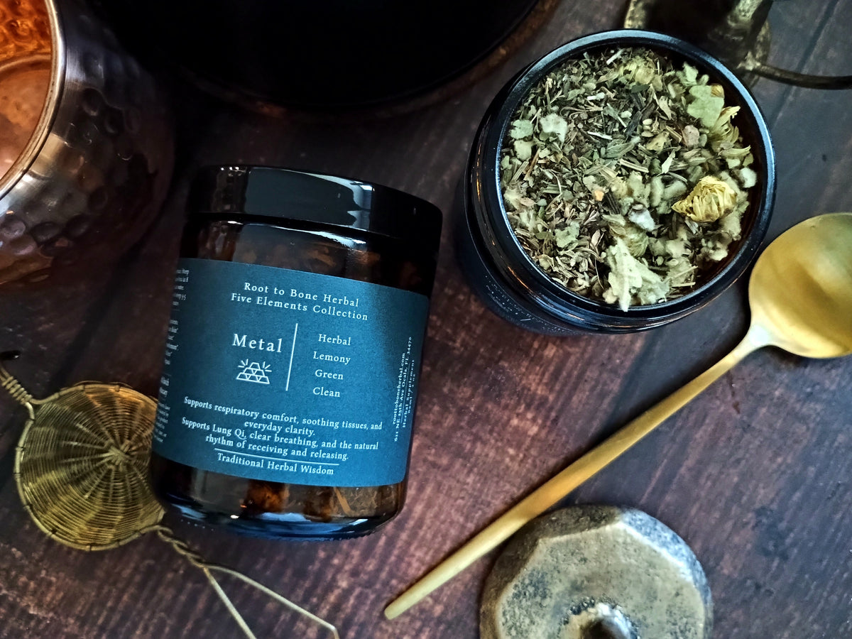 A dark amber glass jar of Metal Root to Bone Herbal Five Elements Collection with a dark blue label, shown next to an open jar of dried herbs and a gold spoon on a dark wooden background.