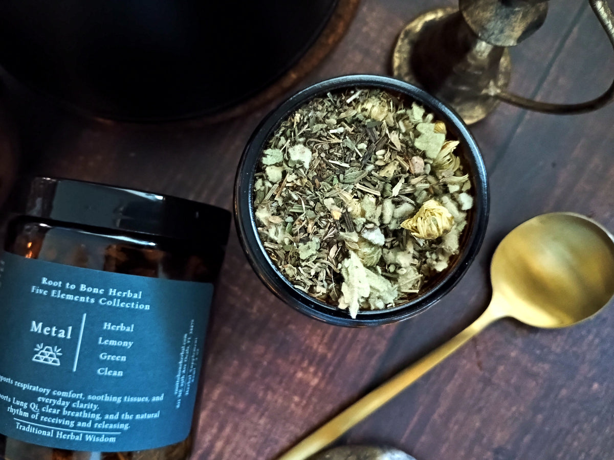 An amber glass jar candle with a dark blue label from the Root to Bone Herbal Five Elements Collection, shown next to a small black bowl filled with dried botanicals and a gold spoon on a dark wooden surface.