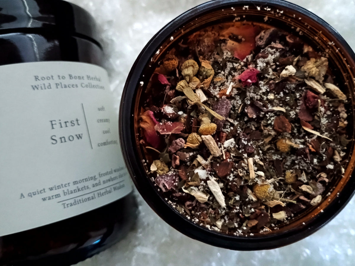 An amber glass jar filled with a dried botanical blend sits in front of a dark bottle labeled 'First Snow'. The jar is photographed from above on a white textured surface, showing the contents of the herbal mixture.