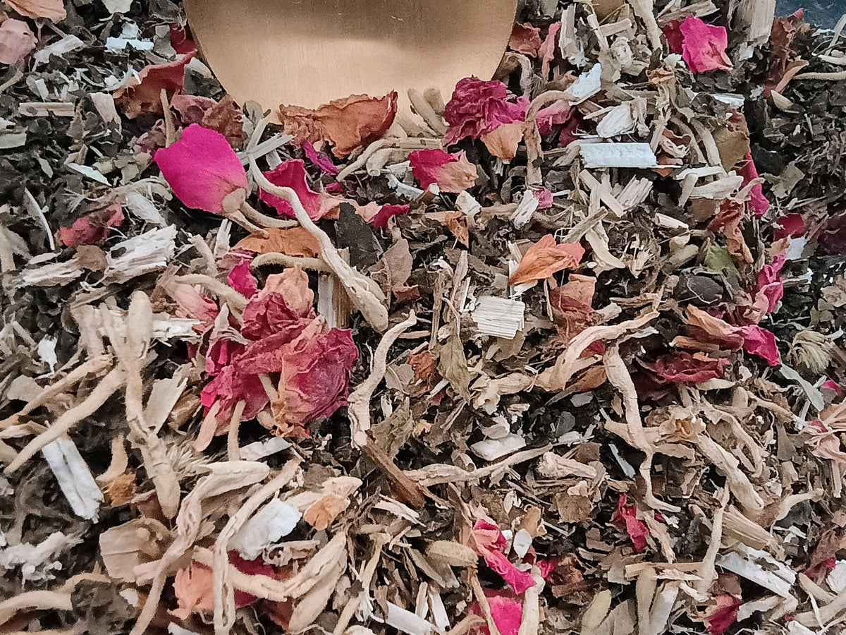 Close-up of loose herbal tea blend with dried rose petals, roots, bark pieces, and small white crystals, with a wooden scoop visible at the top.
