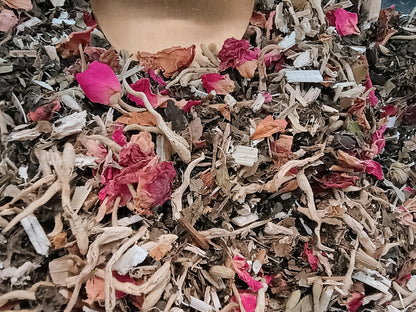 Close-up of loose herbal tea blend with dried rose petals, roots, bark pieces, and small white crystals, with a wooden scoop visible at the top.
