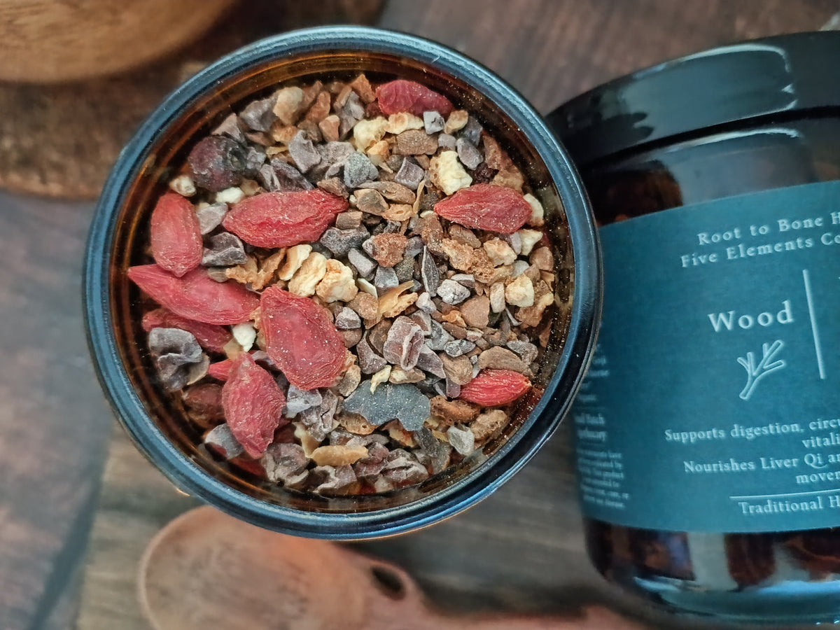An open dark amber jar showing a textured herbal blend with red goji berries, chocolate pieces, and bark, next to the labeled dark blue jar on a rustic wooden surface.