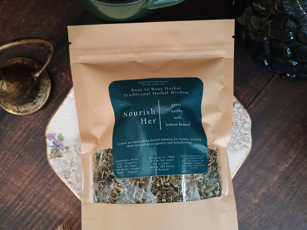 A kraft paper stand-up pouch of Nourish Her herbal tea, featuring a dark teal label, displayed on a decorative floral plate over a dark wooden table.
