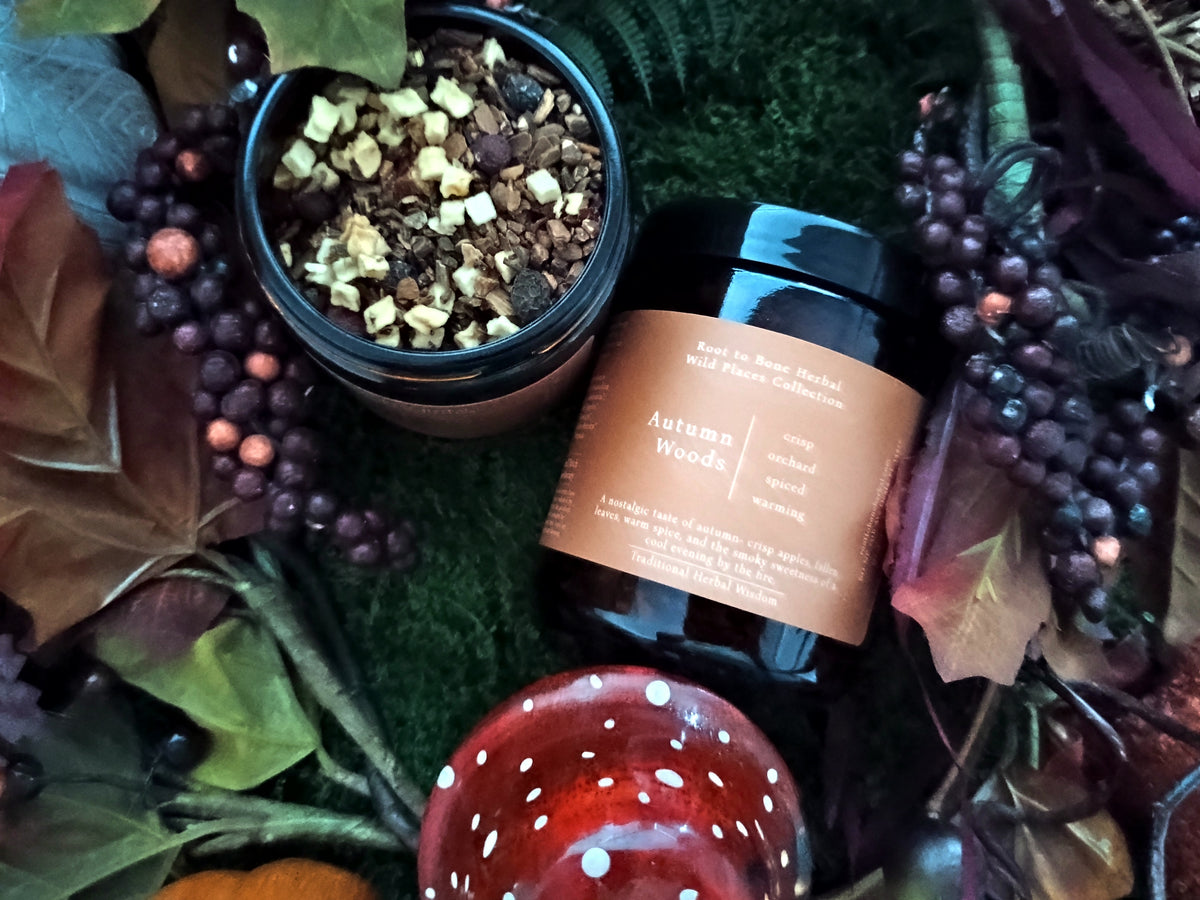 An autumn-themed product flat lay featuring a jar of 'Autumn Woods' herbal tea and an open jar of dried botanicals, styled with moss, fall leaves, berries, and a red polka-dot mushroom.
