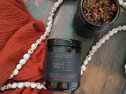 A dark amber glass jar labeled 'Sangria Moon' by Root to Root Herbal, styled on a rustic wooden surface with a rust-red fabric, a beaded necklace, and a second open jar of dried herbs.