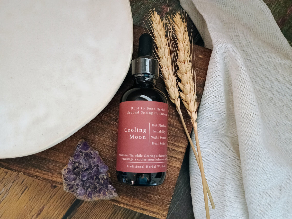 A bottle of Cooling Moon by Root to Bone Herbs, styled on a wooden board with dried wheat stalks, a white ceramic plate, a purple crystal, and a white linen cloth.