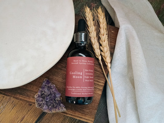A bottle of Cooling Moon by Root to Bone Herbs, styled on a wooden board with dried wheat stalks, a white ceramic plate, a purple crystal, and a white linen cloth.