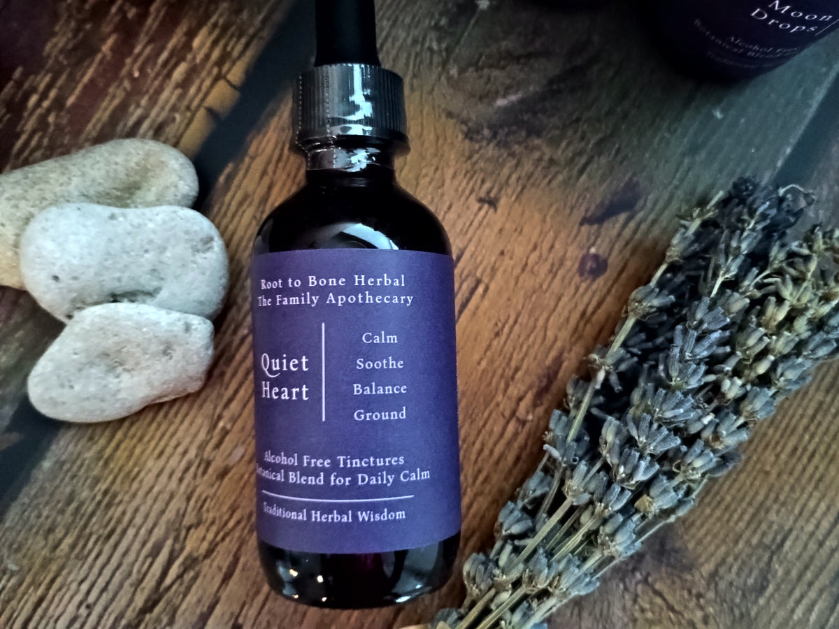A dark amber glass dropper bottle with a purple label reading 'Quiet Heart' by Root to Bone Herbal, styled on a rustic wooden surface with smooth stones and dried lavender.