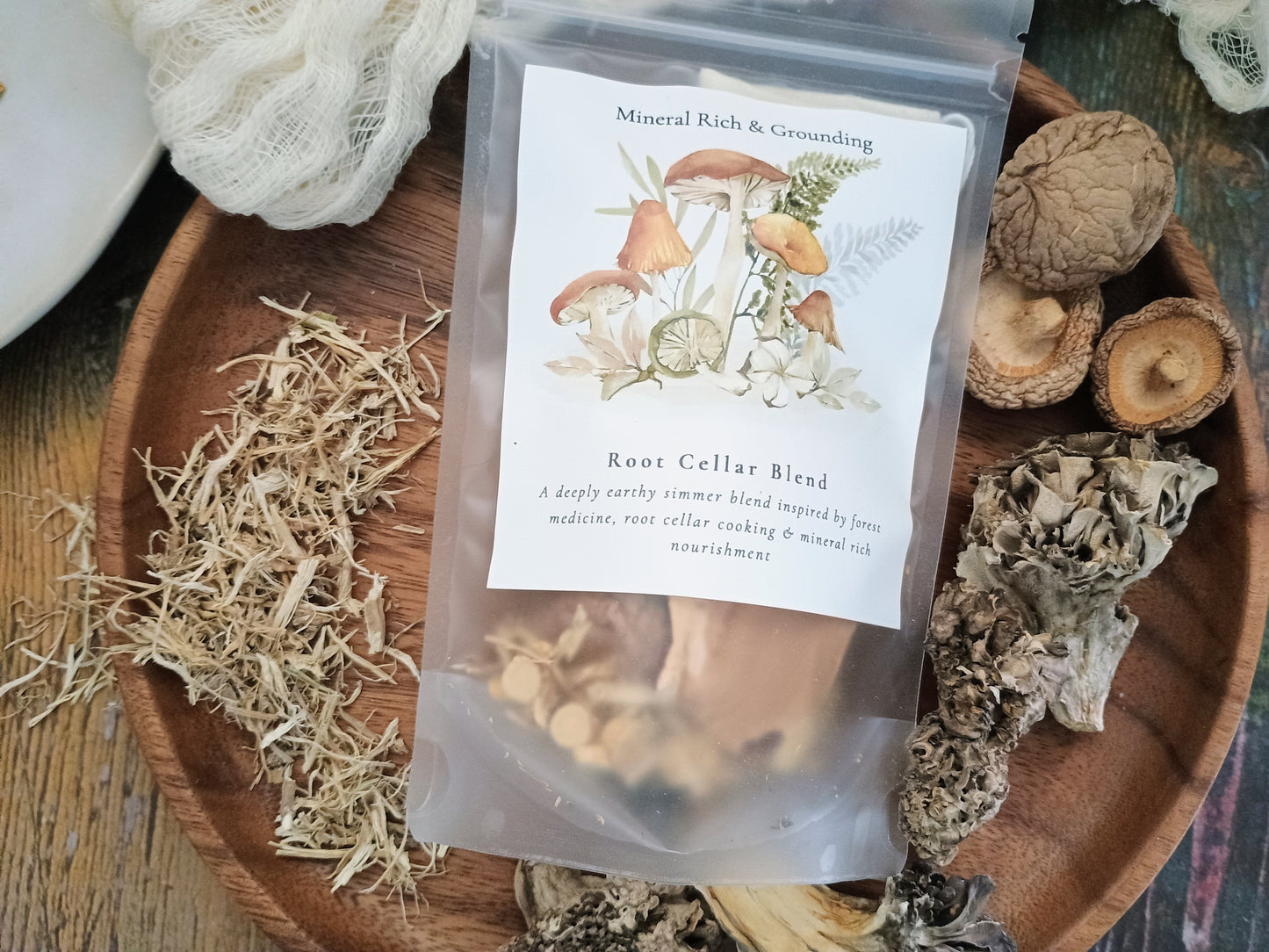 A clear stand-up pouch labeled 'Root Cellar Blend' with mushroom and herb illustrations on the front, placed on a wooden tray surrounded by dried herbs and mushrooms.