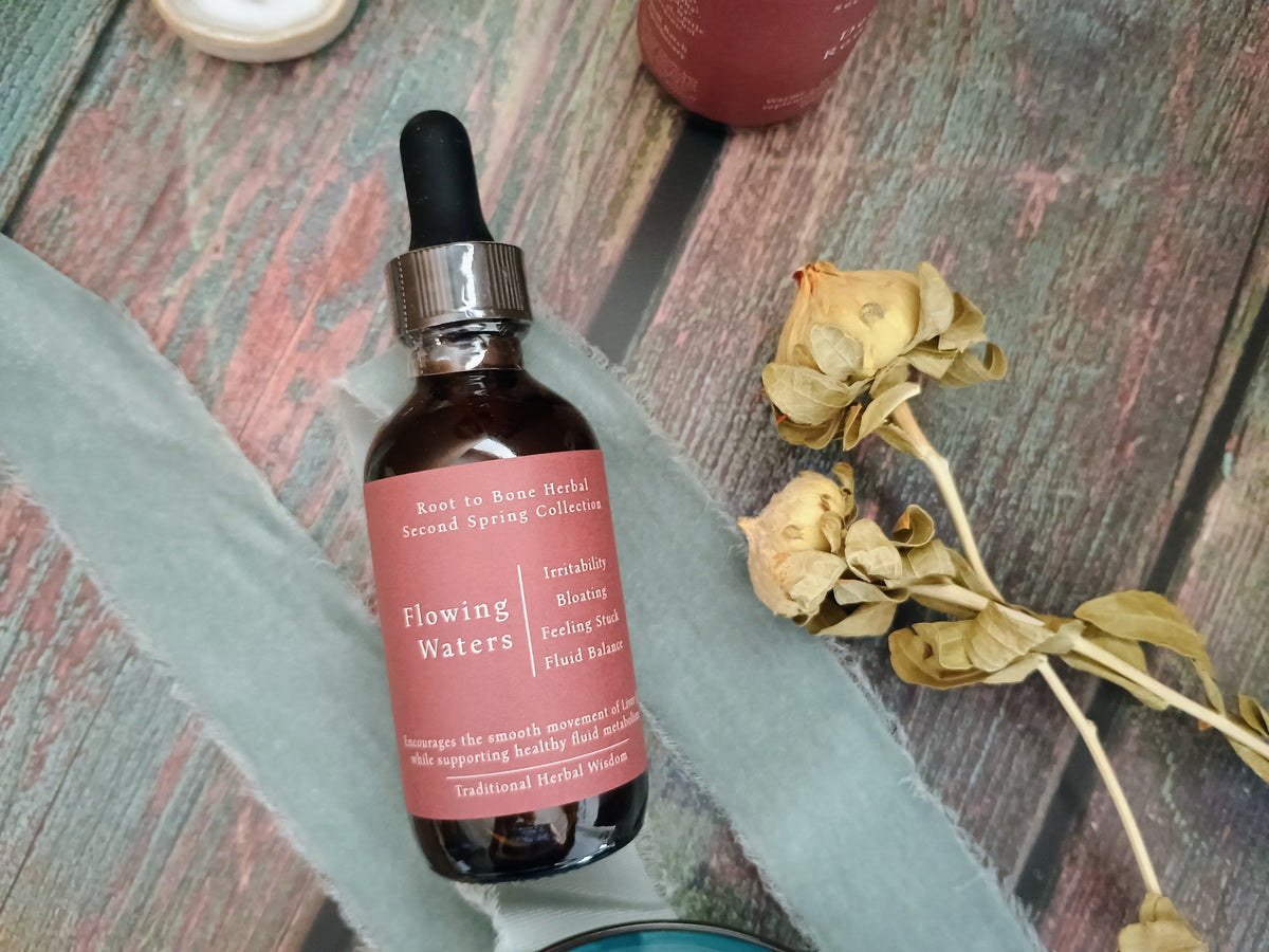A bottle of Flowing Waters herbal with a pink label and dropper cap, styled on a rustic wooden surface with dried flowers and a translucent cloth.