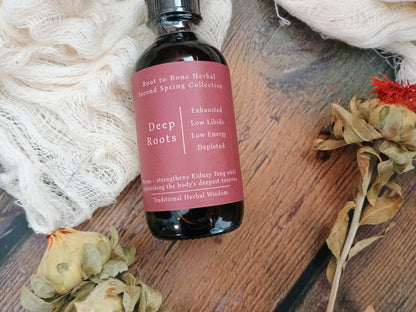A dark glass bottle of herbal tincture labeled 'Deep Roots' by Root to Bone Herbal, styled on a rustic wooden surface with white gauze and dried flowers.