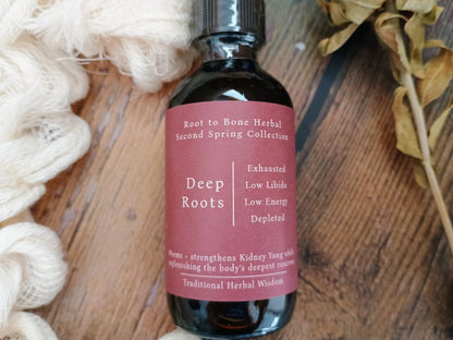 A dark amber glass bottle of Root to Bone Herbal Deep Roots tincture with a dusty rose label, styled on a rustic wooden surface with white wool and dried flowers.