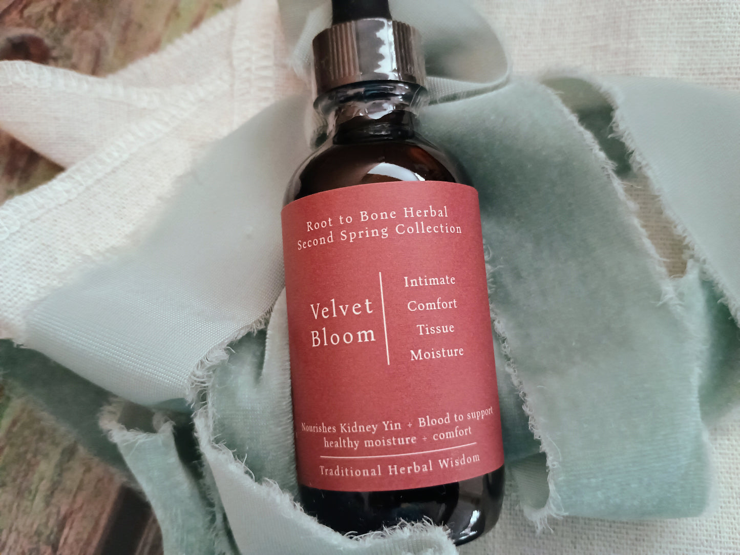 An amber glass dropper bottle labeled 'Velvet Bloom' from the Root to Bone Herbal Second Spring Collection, resting on a light sage green fabric cloth.
