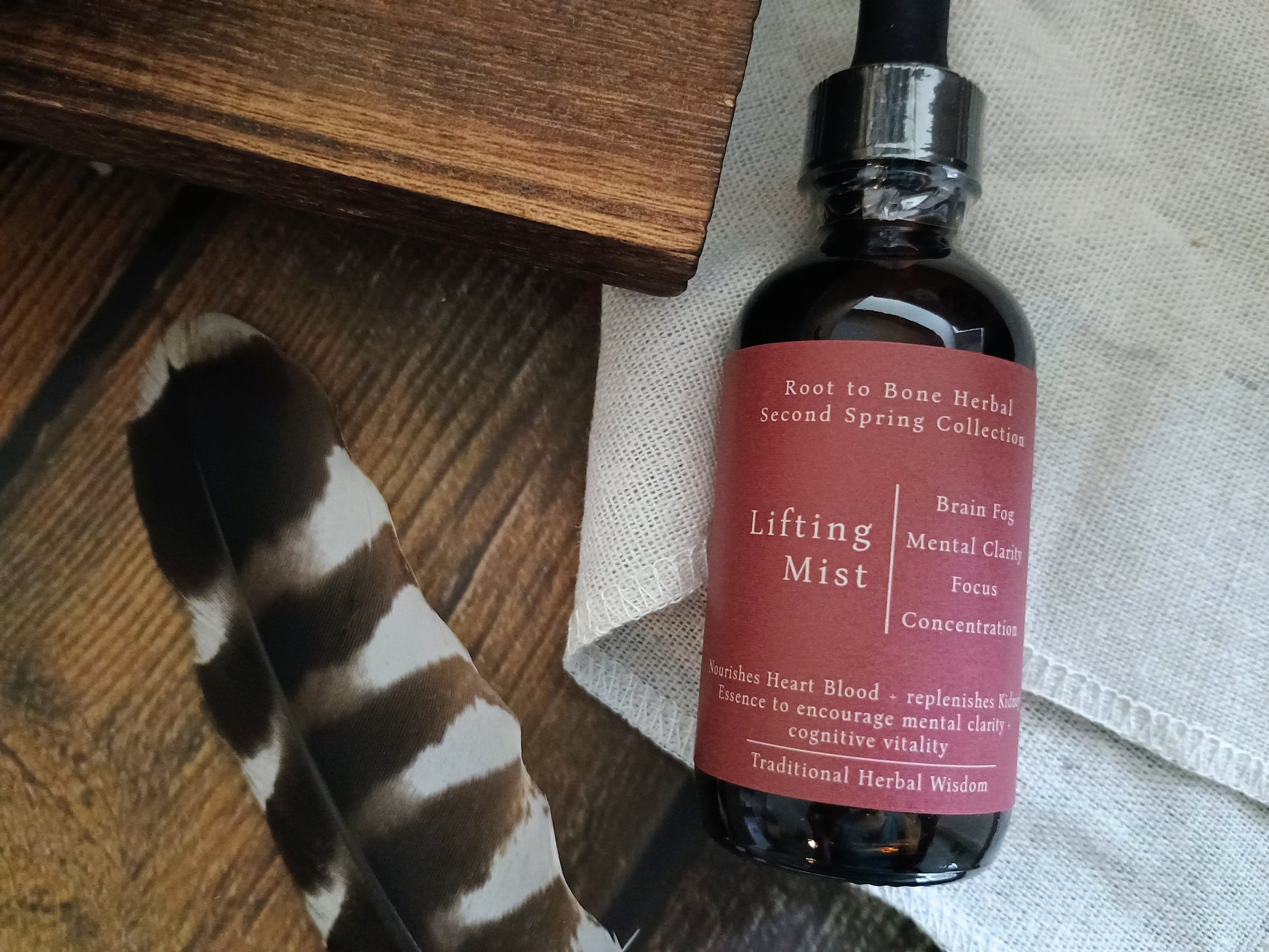 An amber glass dropper bottle labeled 'Lifting Mist' from the Root to Bone Herbal Second Spring Collection, styled on a rustic wooden surface with a black-and-white feather and a light fabric background.