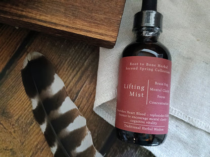 An amber glass dropper bottle labeled 'Lifting Mist' from the Root to Bone Herbal Second Spring Collection, styled on a rustic wooden surface with a black-and-white feather and a light fabric background.