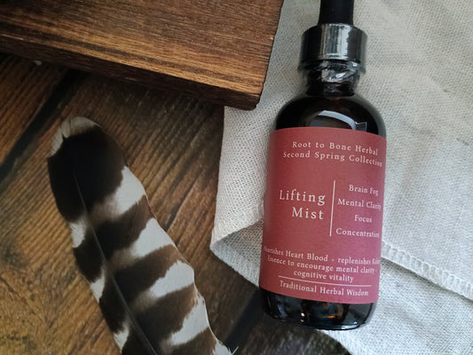 An amber glass dropper bottle labeled 'Lifting Mist' from the Root to Bone Herbal Second Spring Collection, styled on a rustic wooden surface with a black-and-white feather and a light fabric background.