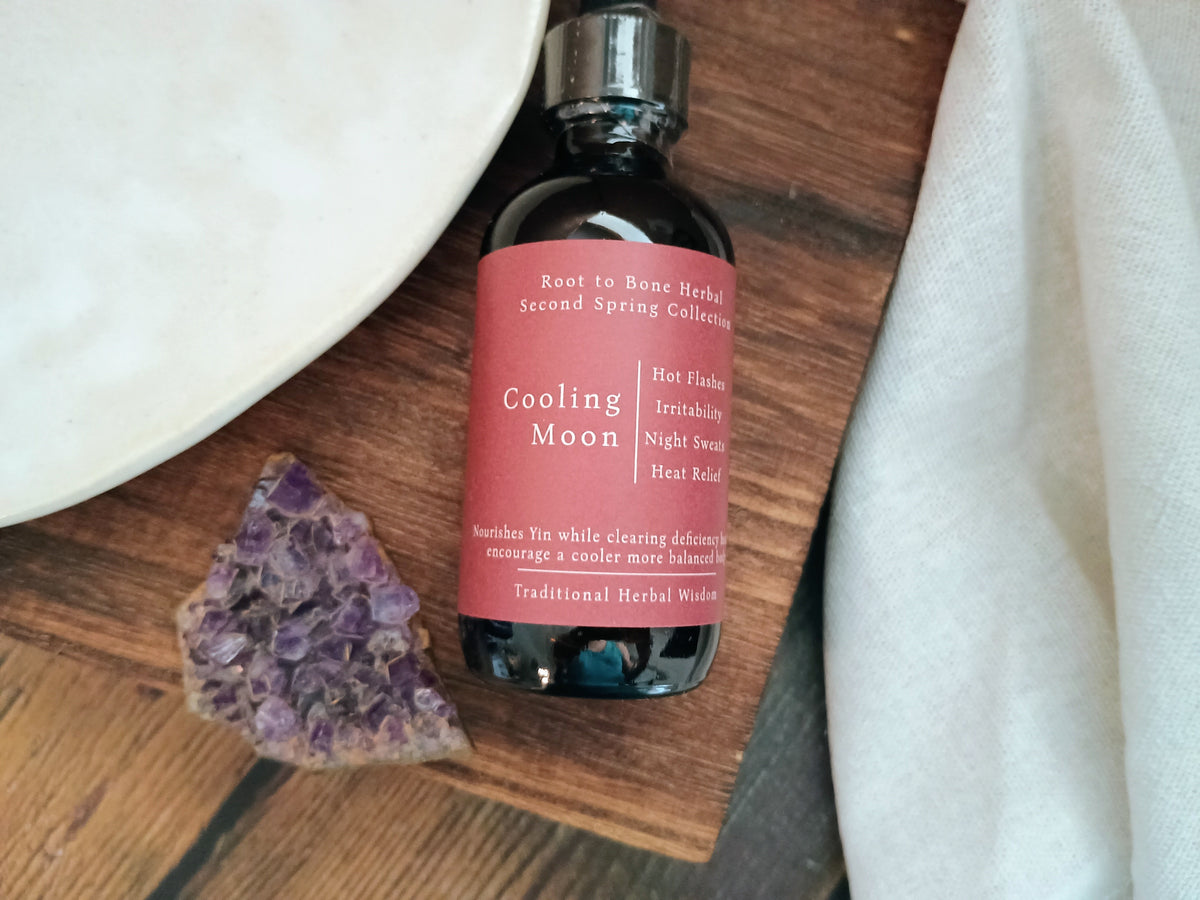 A dark amber glass bottle of Root to Bone Herbs 'Cooling Moon' herbal tincture, featuring a dusty rose pink label, styled on a rustic wooden surface with a white ceramic plate, a purple crystal, and a white cloth.