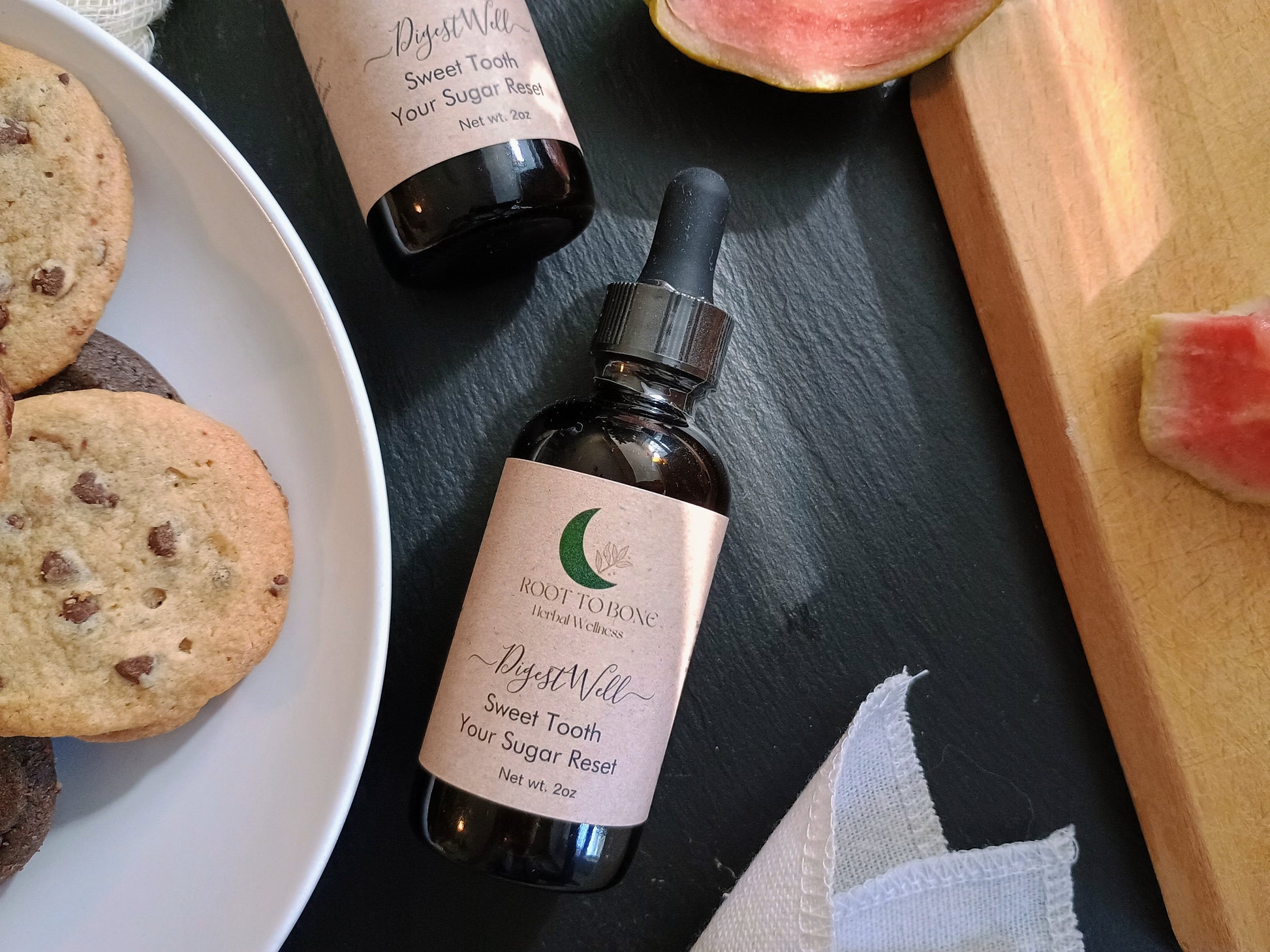 Two bottles of 'Root to Bone' product on a dark surface with cookies and watermelon slices.