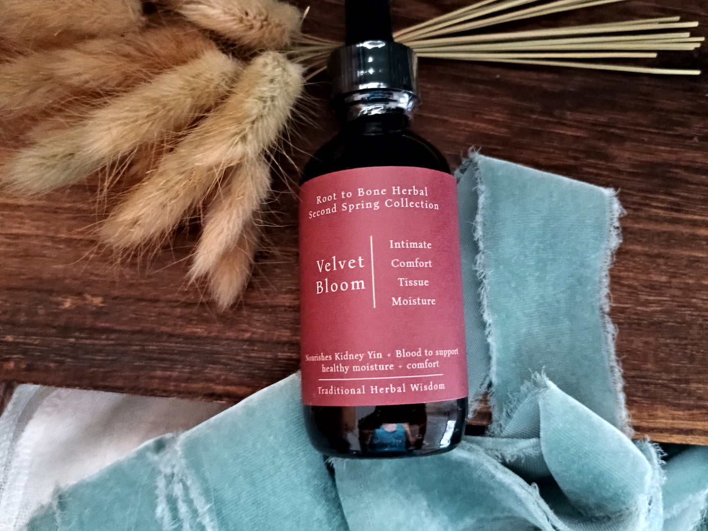 A dark amber glass bottle with a black dropper cap and a dusty pink label reading 'Velvet Bloom' and 'Intimate Comfort Tissue Moisture,' styled on a wooden surface with dried grasses and a light blue cloth.
