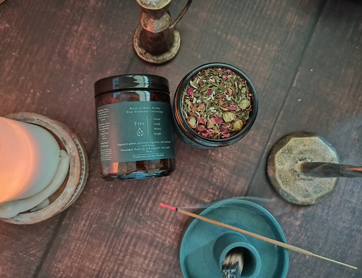 A styled flat-lay of a 'Fire' by Root to Bone Herbal. The dark amber glass jar is shown next to a small open container of dried botanicals, a teal incense holder with a stick, and rustic candles on a dark wooden tabletop.