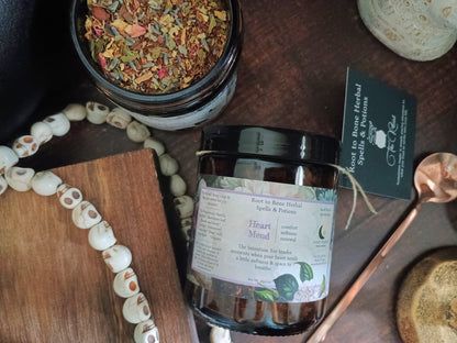 A jar candle labeled 'Heart Mend' by Root to Bone Herbal Spells & potions, displayed on a dark wooden surface with a wooden spoon, a beaded necklace, a small wooden board, and a dried flower jar.