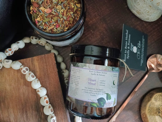 A jar candle labeled 'Heart Mend' by Root to Bone Herbal Spells & potions, displayed on a dark wooden surface with a wooden spoon, a beaded necklace, a small wooden board, and a dried flower jar.