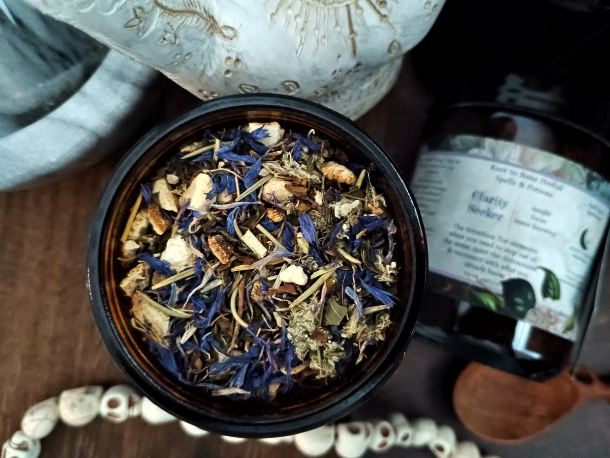 An overhead close-up of a dark bowl filled with loose dried herbal tea blend, featuring blue flowers, green leaves, and tan botanical pieces. A bottle labeled 'Clarity Seeker' sits in the background on a wooden surface, with decorative beads and a ceramic object around it.