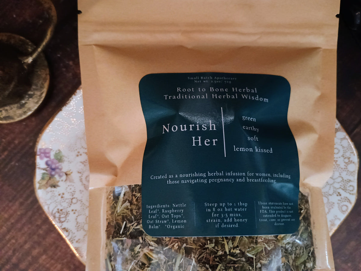A kraft paper pouch of Root to Bone Herbal Wisdom Nourish Her herbal infusion tea, with a dark teal label, shown on a patterned ceramic plate.