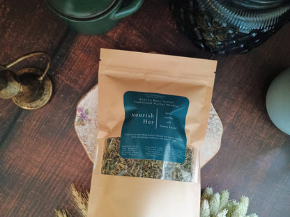 A kraft paper pouch of 'Nourish Her' herbal tea with a dark teal label, displayed on a decorative floral plate on a dark wooden table.
